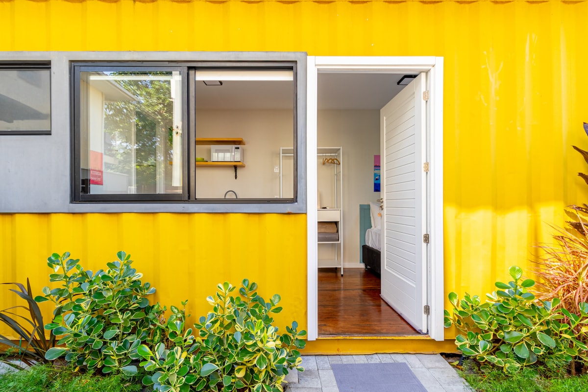 The entrance to a compact unit is framed by vibrant yellow exterior walls. A white door opens into a well-lit interior, showcasing a minimalist space with natural wood flooring. Lush greenery lines the walkway leading to the door, enhancing the inviting atmosphere.