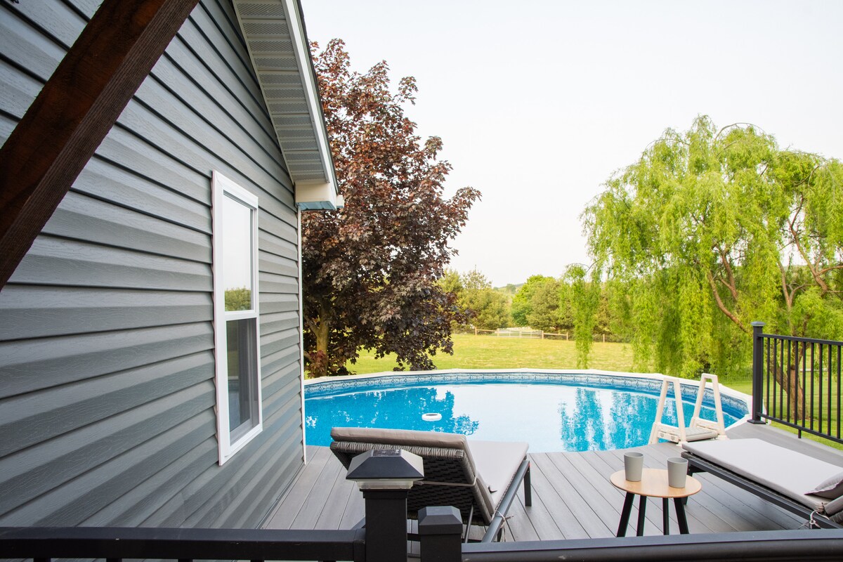 This image features a side view of a newly remodeled home with a gray exterior, showcasing a private pool surrounded by a spacious deck. Comfortable seating is arranged nearby, offering a serene outdoor space with lush greenery in the background.
