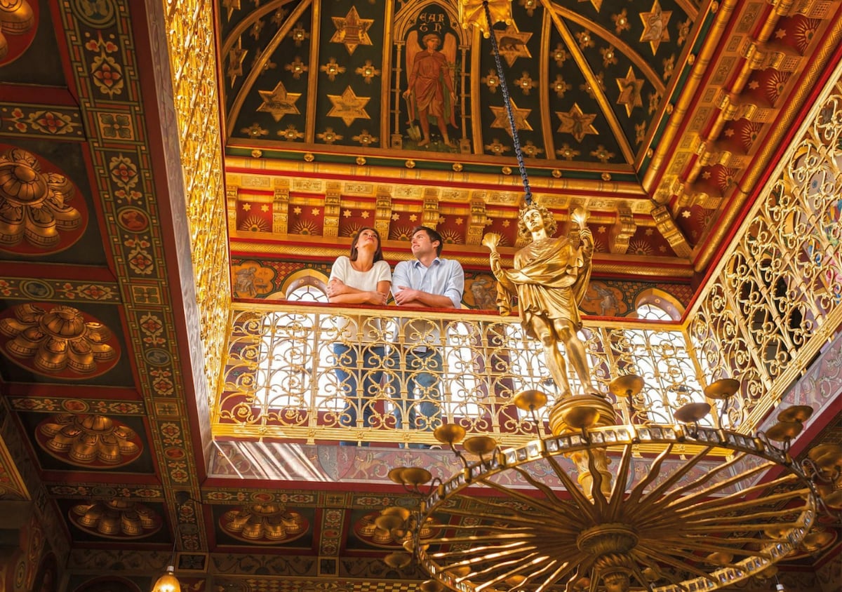 An ornate interior features a richly painted ceiling adorned with gold detailing and star motifs. A decorative balcony with intricate railings overlooks the space, where a golden figure stands prominently. Large, colorfully stained glass windows allow light to illuminate the architectural grandeur.