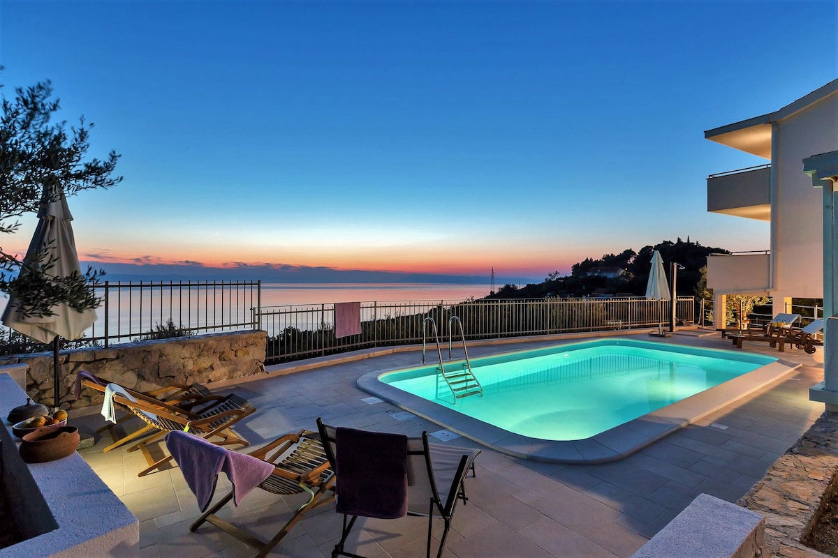A serene outdoor space features a luminous swimming pool surrounded by lounge chairs. The tranquil horizon showcases a gradient of sunset colors, and two umbrellas are positioned for shade. The area is enclosed by a low stone wall, enhancing privacy and the stunning sea view.