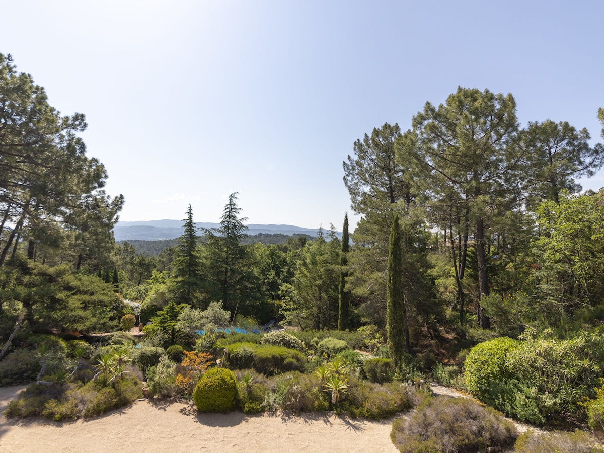 A lush, green landscape is featured, showcasing a variety of trees and shrubs. The image captures a scenic view with gentle hills in the background, extending into the clear blue sky. The natural flora creates a serene and inviting outdoor environment.