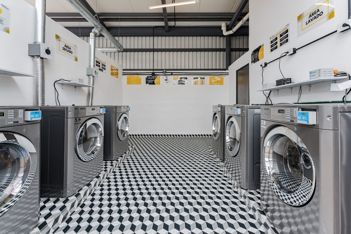 A modern laundry facility is equipped with multiple washing machines and dryers arranged in a spacious layout. The geometric patterned floor enhances the contemporary design. Bright lighting illuminates the area, and clear signage is visible on the walls, indicating various washing options and instructions.