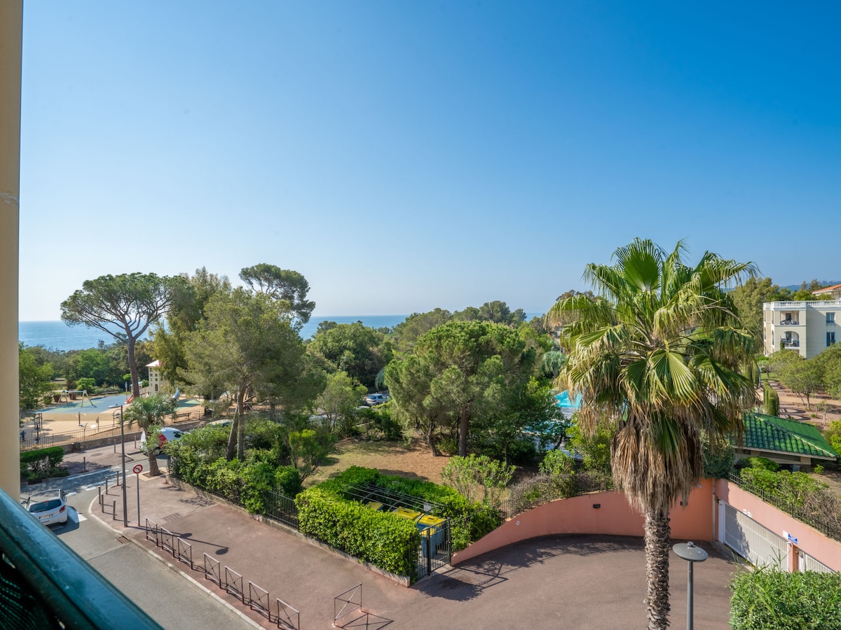A view from the balcony displays a lush landscape featuring palm trees and greenery. In the distance, the sea is visible beyond a park area. The clear blue sky enhances the tranquil setting.
