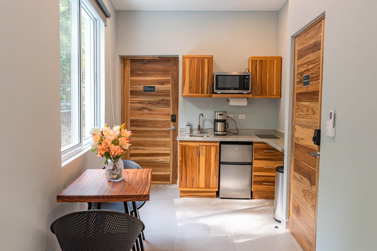 A well-organized kitchenette is visible, featuring wooden cabinetry and essential appliances including a microwave and coffee maker. A small wooden dining table with two black chairs is positioned nearby, complemented by a floral arrangement. Natural light enters through a large window.