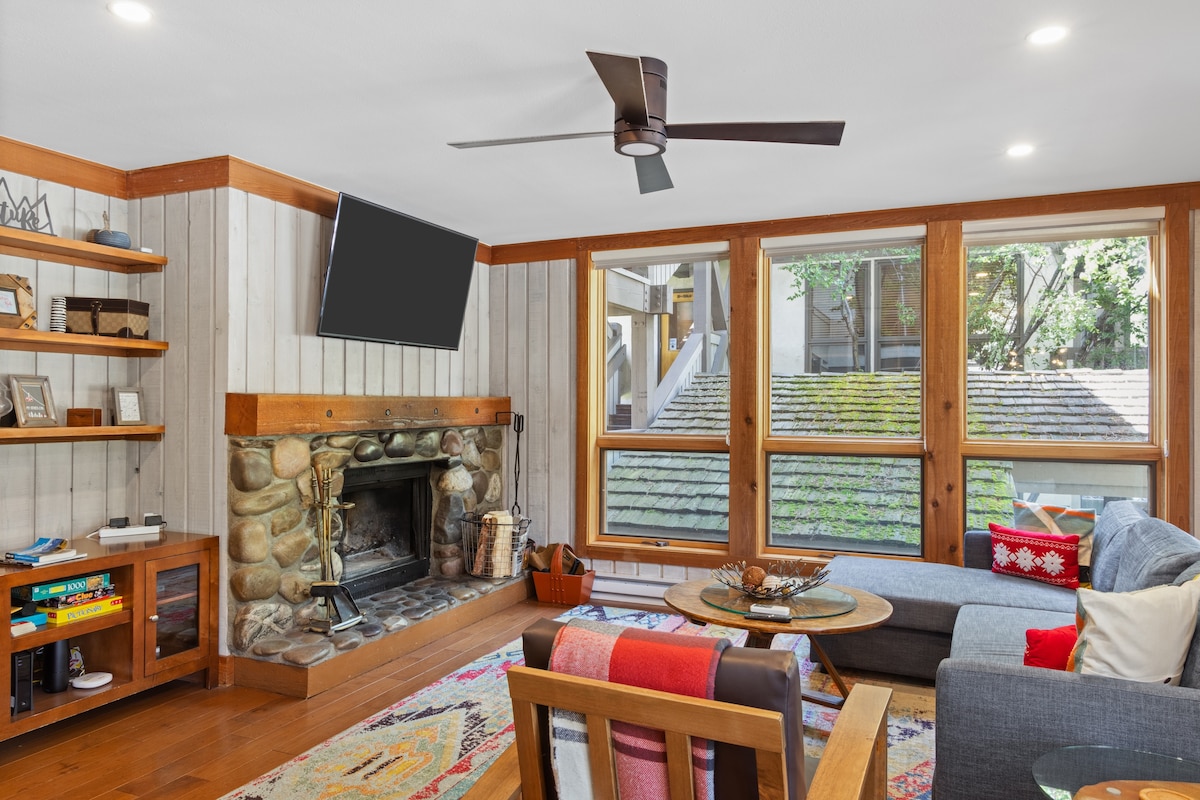 A welcoming living area features a stone fireplace as the focal point, flanked by large windows that invite natural light. A comfortable couch and colorful chairs surround a circular table, while shelves display books and decorative objects, creating a cozy atmosphere.