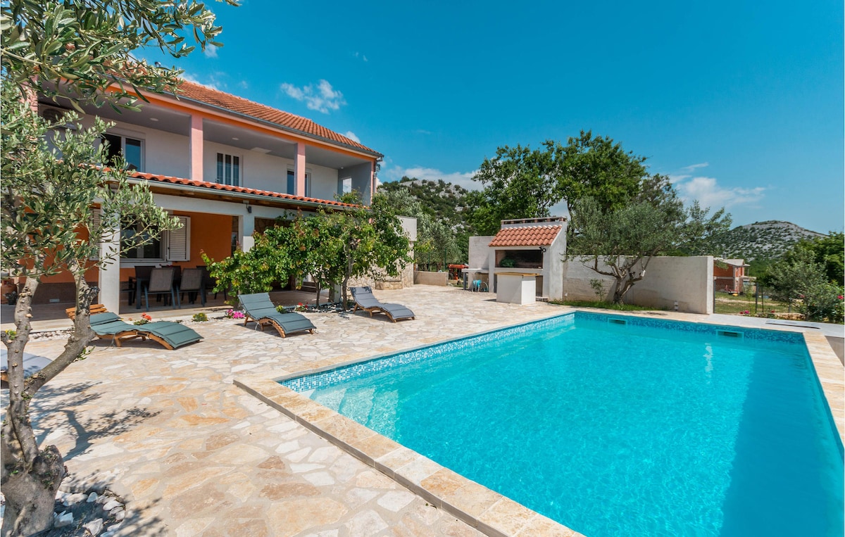 An outdoor private pool is set within a paved area, surrounded by lounge chairs. The large building showcases a balcony with sliding doors, and green trees provide shade. A clear blue sky enhances the inviting atmosphere of this relaxing outdoor space.