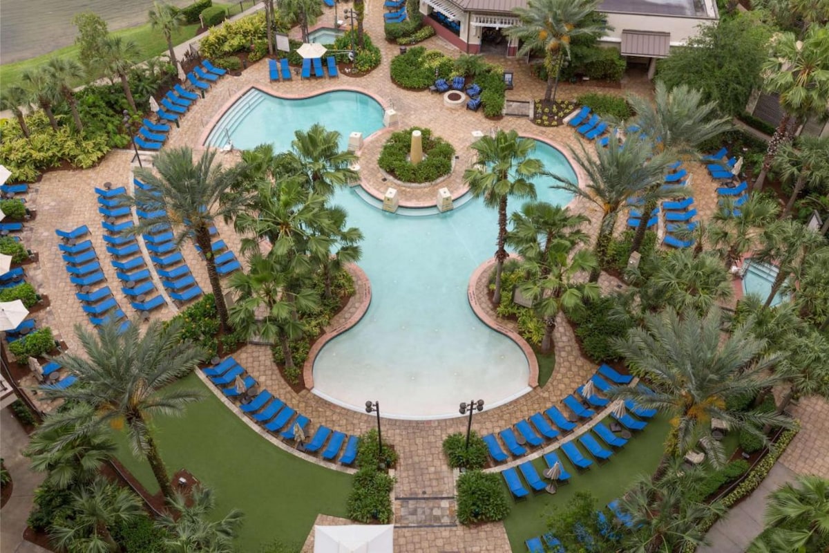 An aerial view of a lagoon-style pool surrounded by lush greenery and palm trees. Vibrant blue lounge chairs line the pool’s edge, offering ample relaxation space. Pathways leading to additional seating areas and water features are visible, creating a serene outdoor environment.