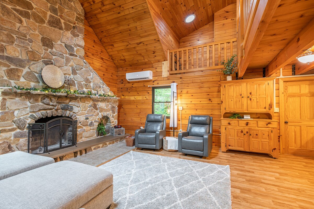 The living area features a vaulted wooden ceiling with exposed beams and a stone fireplace as a focal point. Two leather armchairs are positioned for comfort, accompanied by a large area rug. Natural light filters through the windows, illuminating the warm wooden interior.
