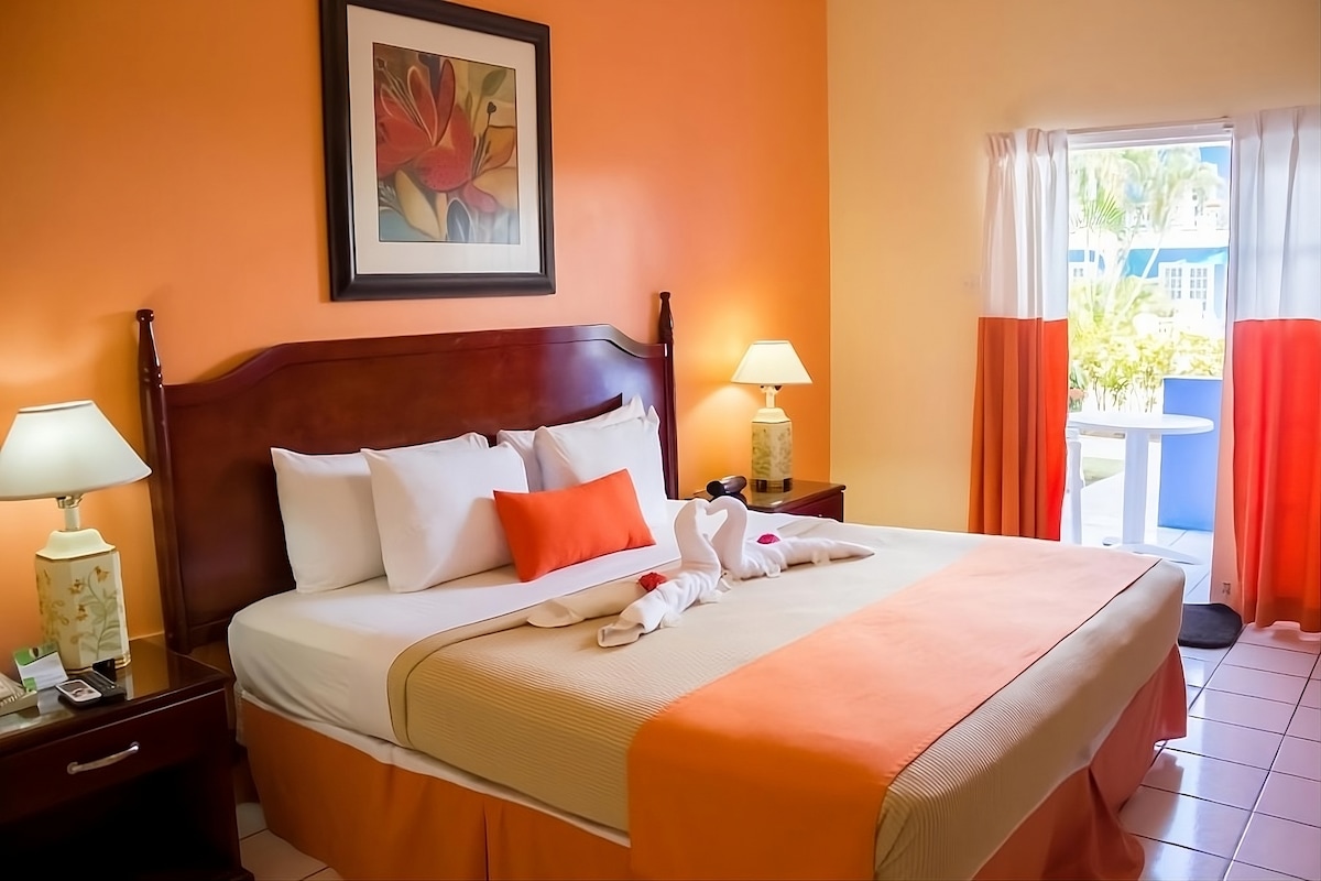 A cozy hotel room features a king bed adorned with crisp linens and vibrant orange accents. Two bedside lamps illuminate the space, and a colorful floral artwork decorates the wall. A sliding door leads to a balcony, inviting natural light into the room.