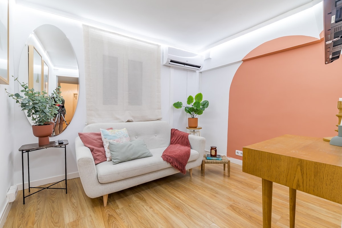 A modern living area features a light-colored sofa adorned with colorful cushions. A small table stands alongside, complemented by a potted plant. The wall displays a warm peach accent, while ample natural light enters through a sheer window covering.