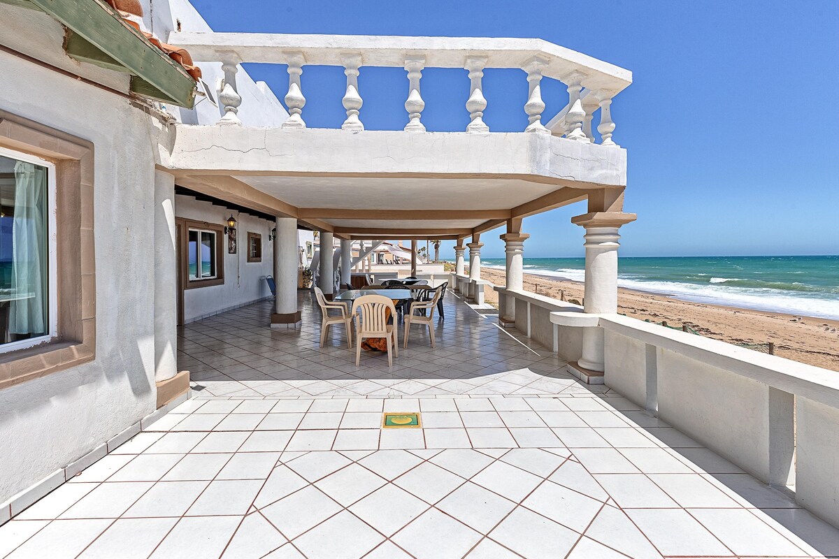A spacious outdoor patio area is featured, showcasing a tiled floor and a stunning view of the beach and ocean. White columns provide architectural interest, while a table and chairs offer a comfortable setting for relaxation or dining. The sky is clear and blue.