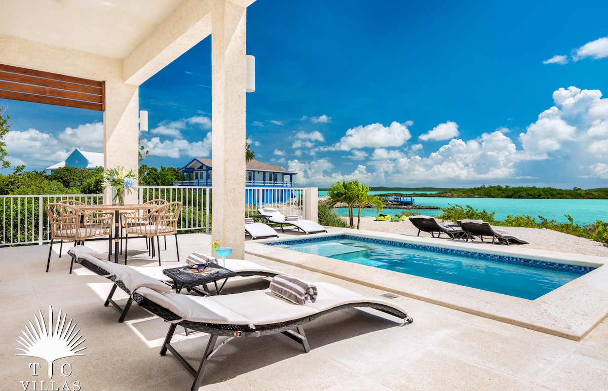 An outdoor terrace features a private infinity pool and lounge chairs, overlooking the serene blue waters of Chalk Sound. A dining area is set with a table and chairs nearby. Lush greenery and bright clouds complement the tranquil environment.