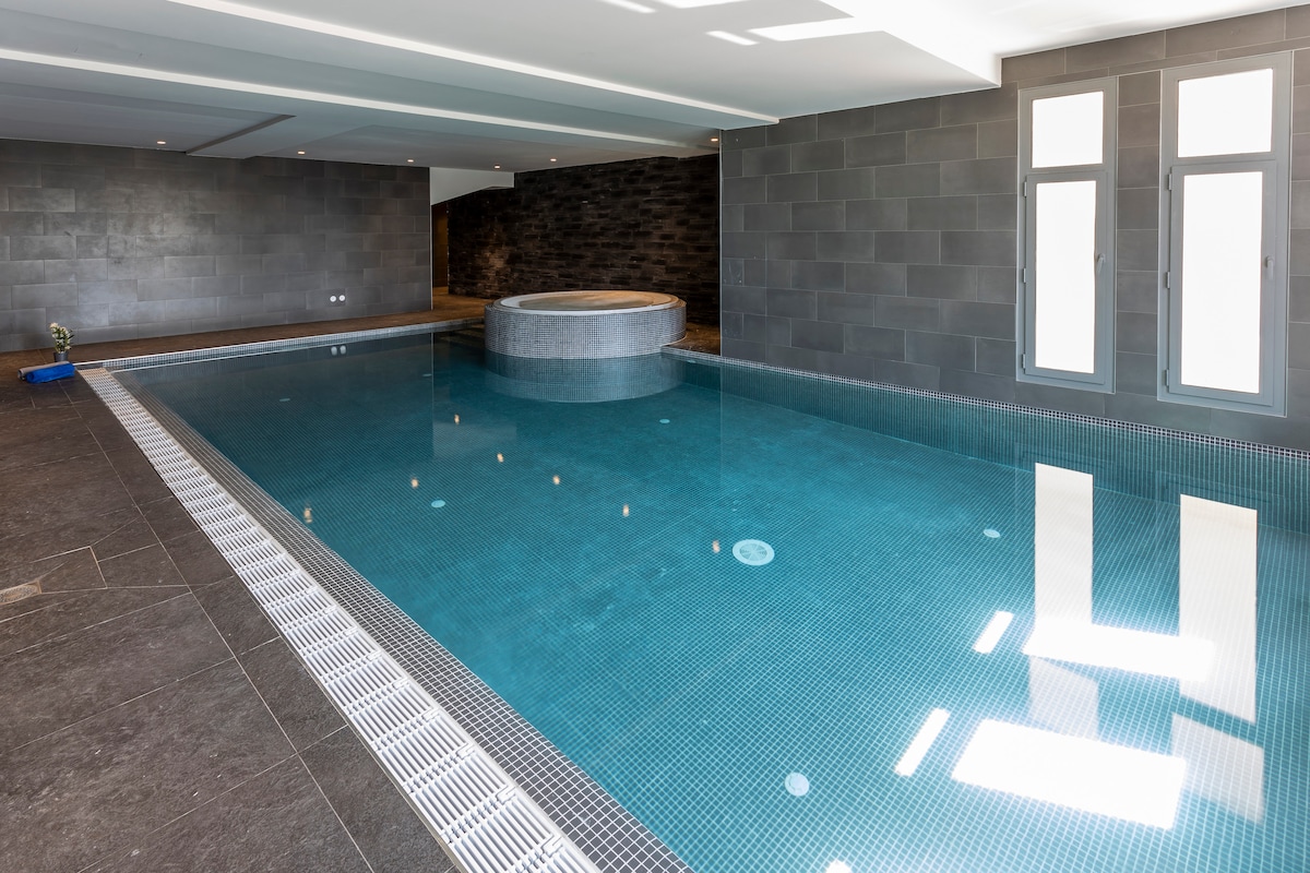 An indoor pool area is showcased, featuring a large, rectangular pool with clear blue water and a circular jacuzzi at one end. The surrounding walls are finished in dark stone, and natural light streams in through several large windows, illuminating the tranquil space.