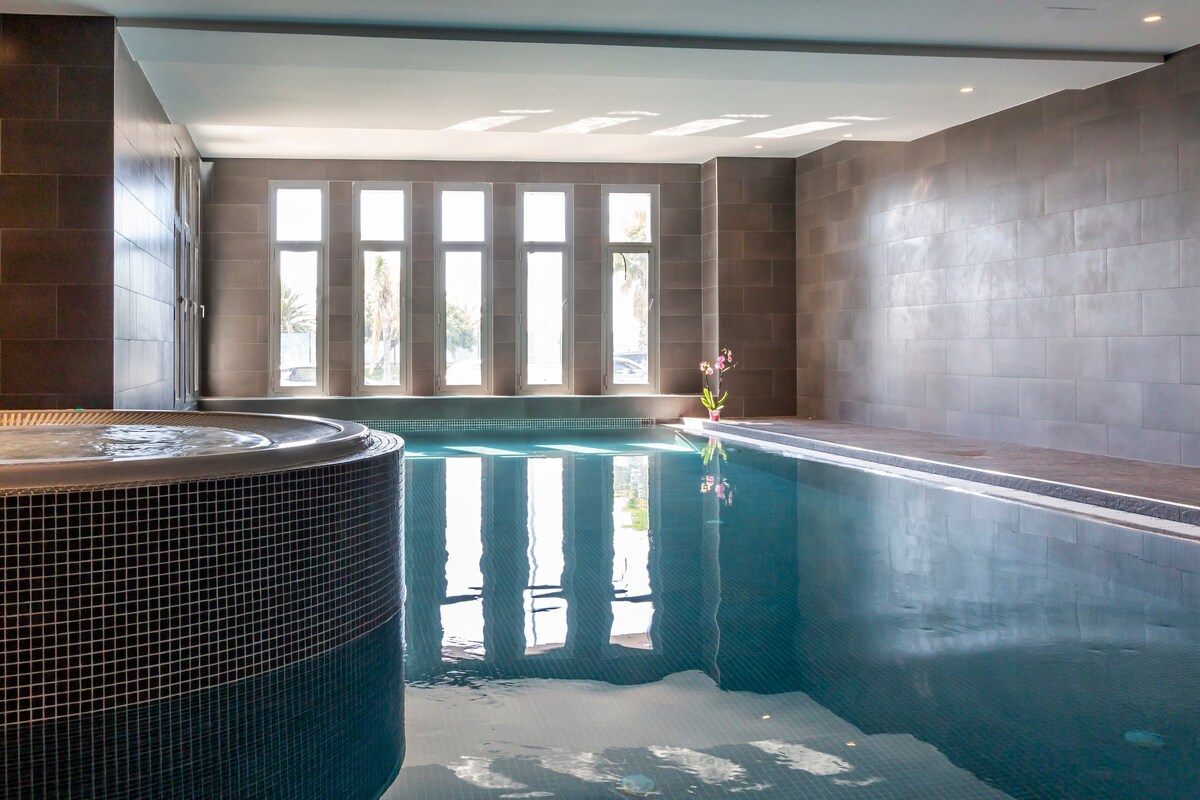 An indoor swimming pool is illuminated by natural light flooding through large windows. The serene water reflects the surrounding walls, complemented by a whirlpool at one end and a planted centerpiece adding a touch of greenery.