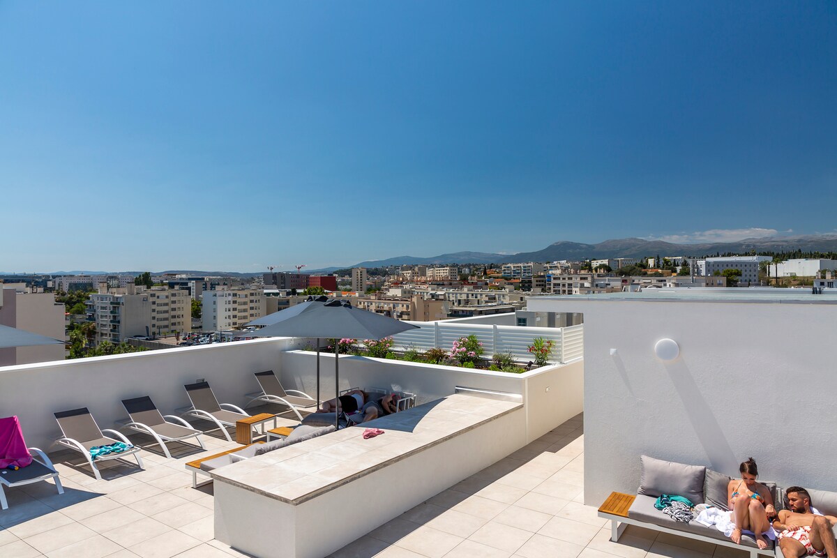 The rooftop pool area features a spacious deck with lounge chairs and a shaded umbrella. Surrounding views of the city and distant hills complete the vibrant atmosphere. A modern design is evident in the clean lines and bright surfaces, creating a welcoming space for relaxation.