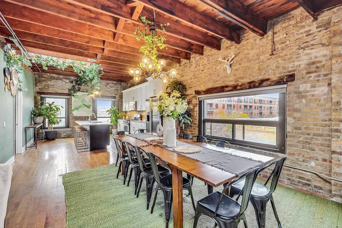 A spacious dining area features a long wooden table surrounded by black metal chairs. Exposed brick walls and wooden beams add warmth to the room. Large windows invite natural light, showcasing greenery and the well-appointed kitchen in the background.