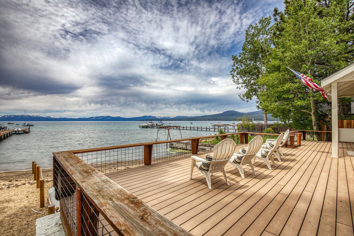 A spacious deck overlooks the tranquil waters of Lake Tahoe, furnished with white lounge chairs. The scene includes a sandy beach extending to the water's edge and boats anchored in the distance, surrounded by lush trees under a cloudy sky.