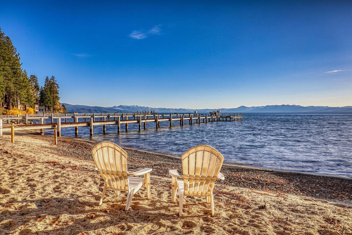 Two white Adirondack chairs are positioned on a sandy beach, facing the gentle waves of Lake Tahoe. A wooden pier extends into the serene water, with distant mountains visible under a clear blue sky.