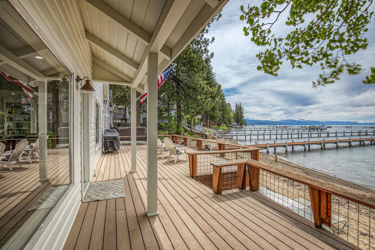 An expansive wooden deck overlooks a private sandy beach and the lake, with several white lounge chairs positioned for relaxation. Reflections of the nearby trees and the lake can be seen in the large glass doors. The serene setting invites outdoor enjoyment.