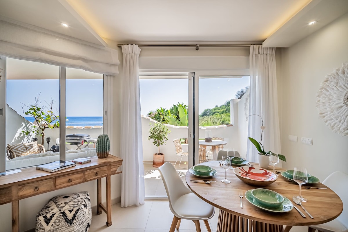 A dining area is presented with a circular wooden table set for meals, accompanied by modern white chairs. Large glass doors offer a view of the outdoor terrace and ocean beyond, while light curtains frame the scene. Decorative elements include green tableware and a plant in the background.