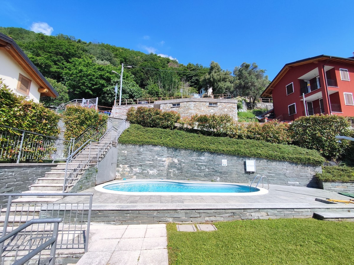 A kidney-shaped swimming pool is set in a landscaped area, surrounded by neatly trimmed hedges and greenery. Stone steps lead from the pool area to a hillside that is partly bordered with residences, showcasing a pleasant blend of nature and architecture under a clear blue sky.