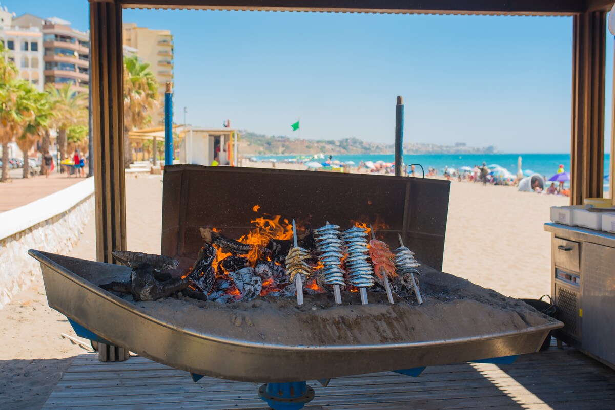 A beachside grill is shown with flames and skewers of food cooking over the open fire. The sandy beach is visible in the background, with people enjoying the sun and sea, and palm trees lining the walkway.