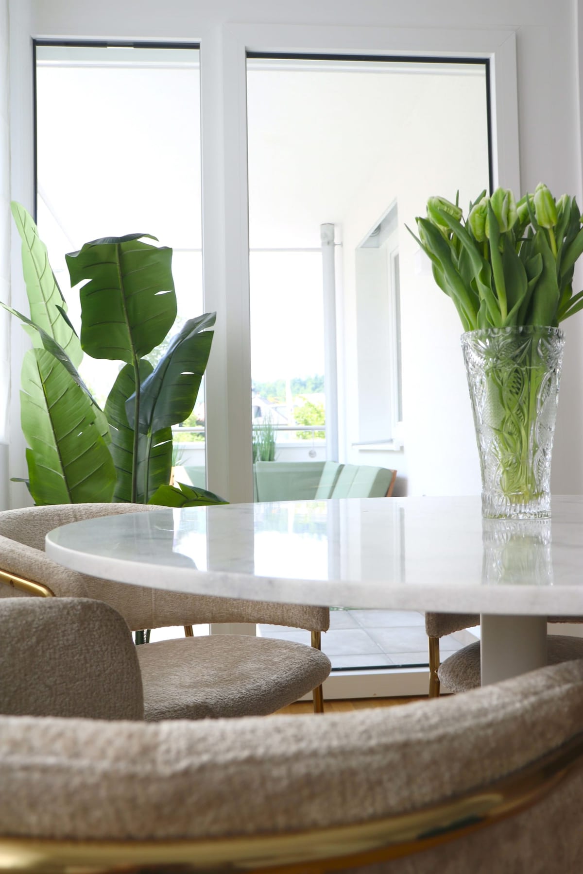 A light-filled dining area features a round marble table surrounded by plush chairs. In the background, green plants can be seen beside large glass doors that lead to a balcony, enhancing the freshness of the space.