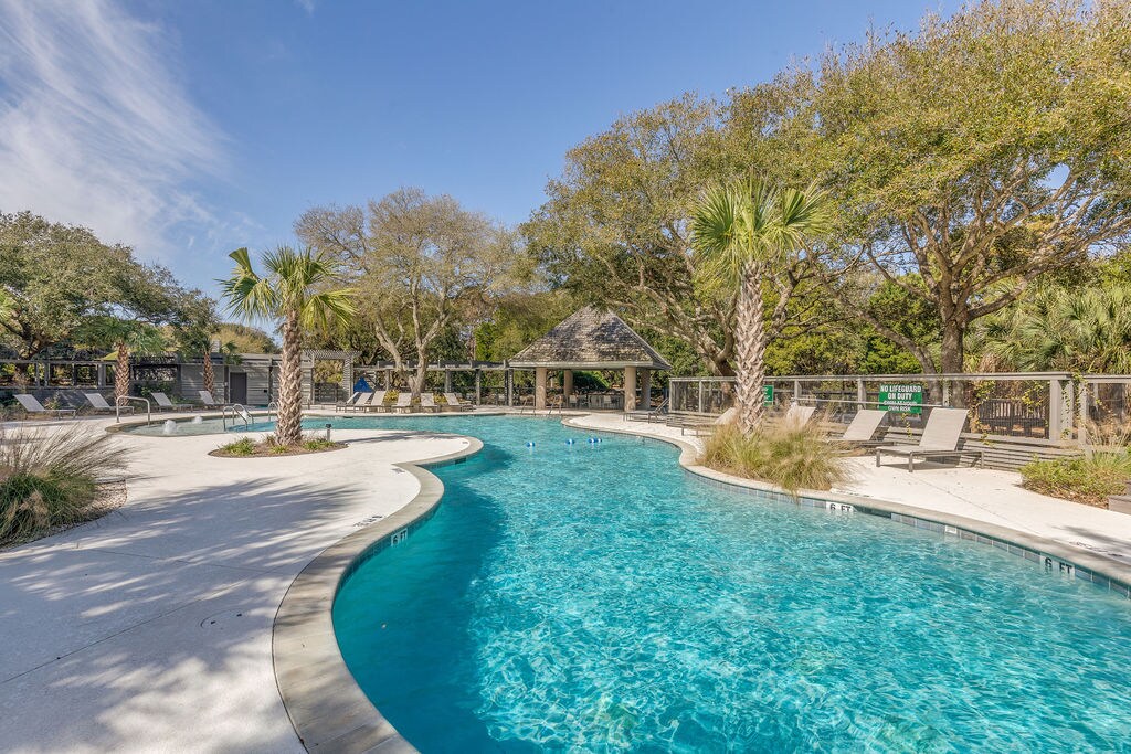 The community pool area features a winding design surrounded by lush landscaping and palm trees. Loungers are positioned around the pool, and a shaded pavilion is visible in the background, offering an inviting space for relaxation.