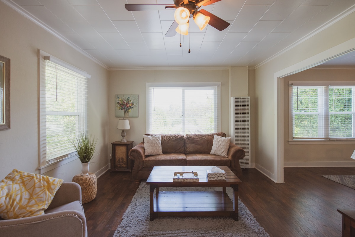 A cozy living area is depicted, featuring a comfortable brown sofa and a wooden coffee table. Light streams through large windows adorned with white blinds, illuminating the space. A corner lamp and greenery add to the inviting setting.
