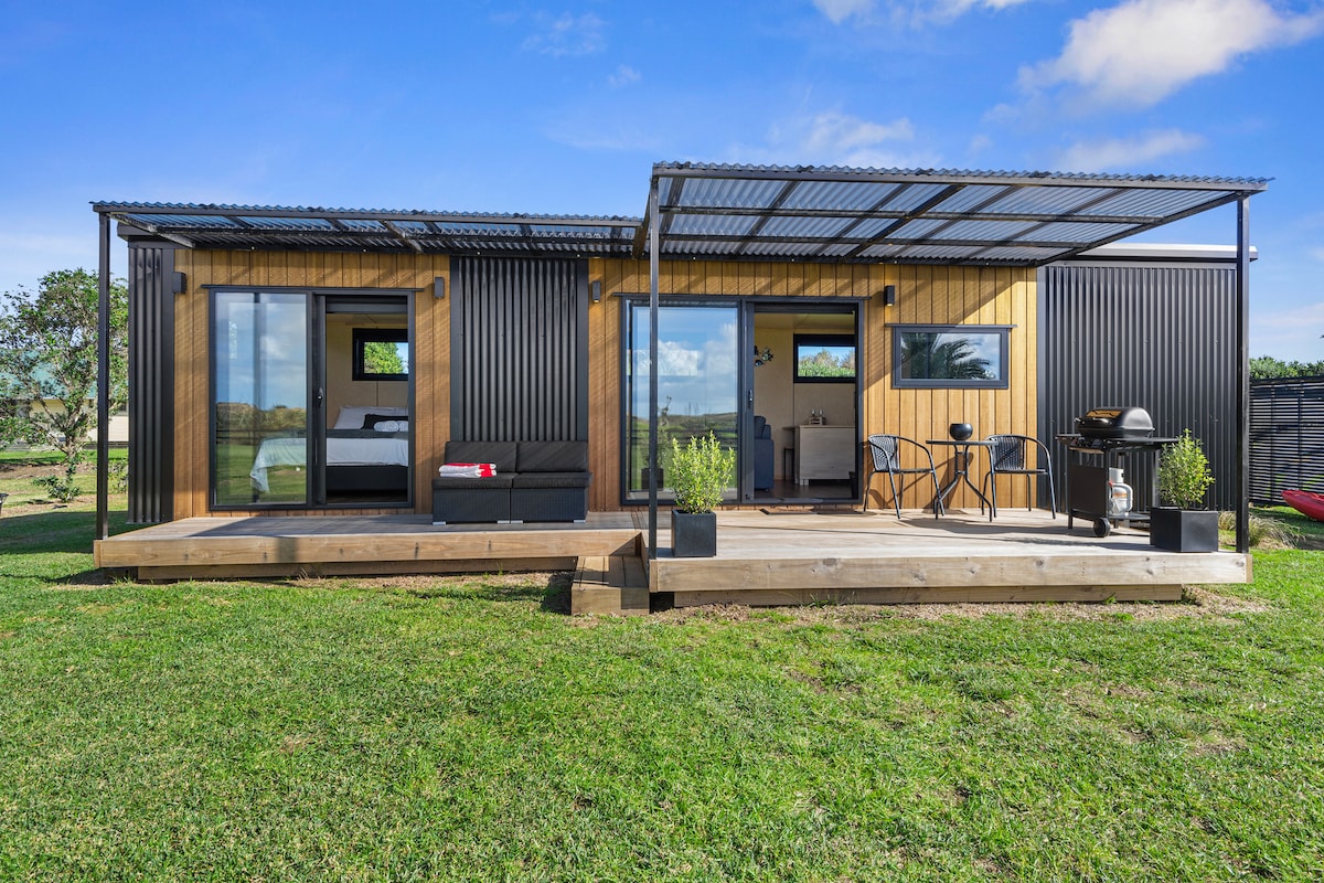 A modern cottage exterior features a sleek combination of wood and metal materials. A covered deck provides a shaded area with seating and a barbecue, inviting outdoor enjoyment. Large glass doors offer views of the surrounding greenery and easy access to the indoor living space.