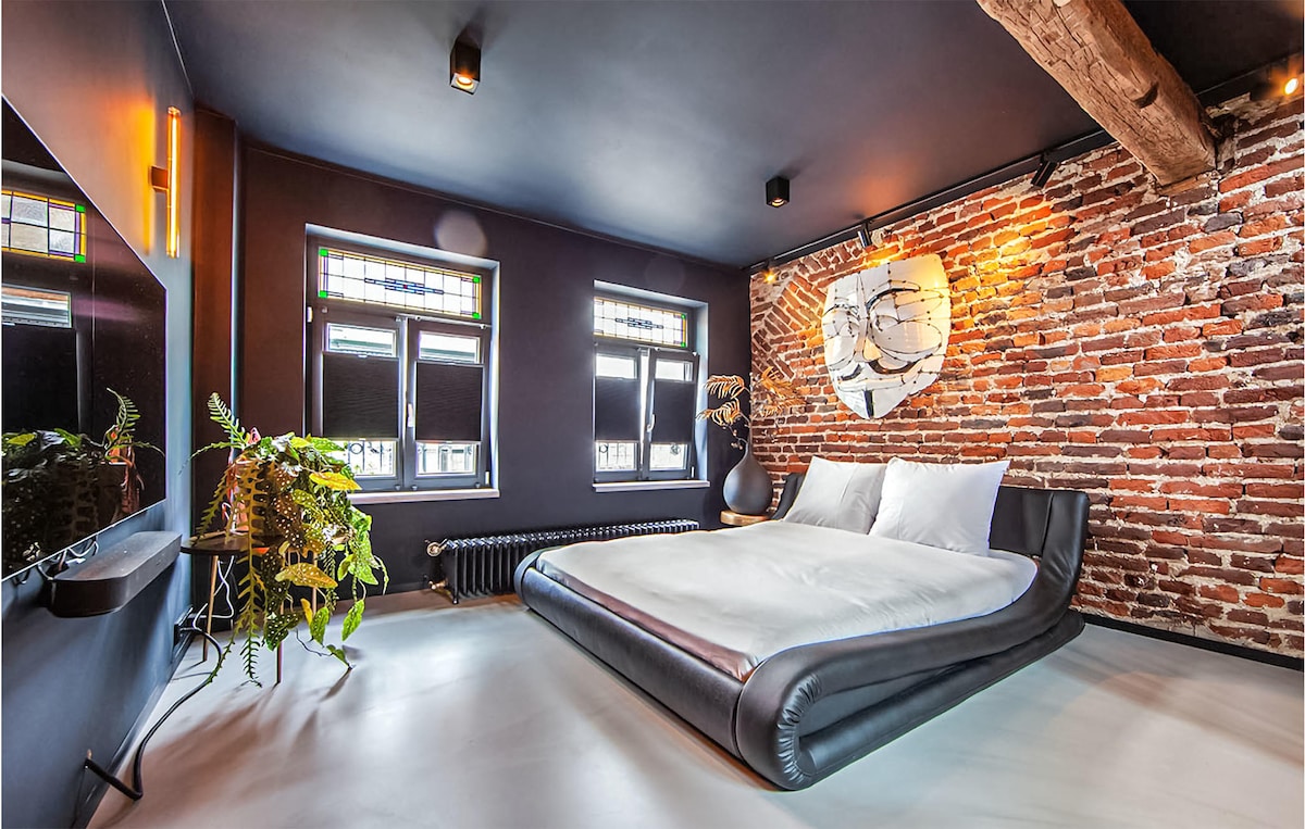 A stylish bedroom features a spacious bed with white linens against a backdrop of exposed brick walls. Natural light enters through windows adorned with black blinds. A sleek television is mounted on the wall, and a potted plant adds a touch of greenery to the space.