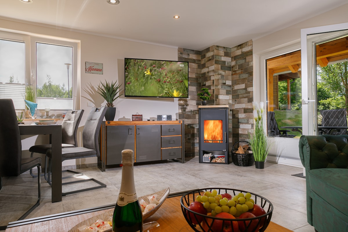 A living and dining area features a wood-burning fireplace and a flat screen satellite TV. Large windows allow natural light, and a dining table with chairs complements the space. A decorative bowl with fresh fruit sits on a coffee table, adding to the welcoming ambiance.