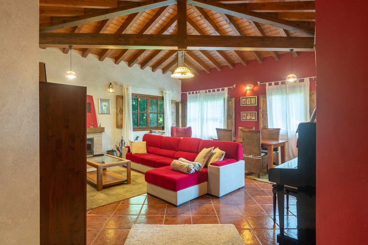 A spacious living area features a red sectional sofa arranged around a coffee table on tiled flooring. High wooden beams enhance the open feel, with natural light filtering through large windows dressed in light drapes. A dining area is also visible, along with a piano.