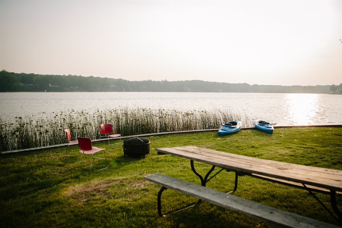 A serene lakeside view showcases two kayaks resting on the grass, accompanied by a picnic table and a fire pit nearby. Colorful chairs are positioned close to the water's edge, with tall grass framing the scene against the glistening surface of the lake.