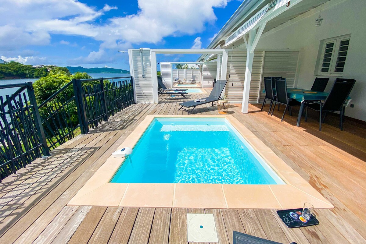 A private terrace features a small pool surrounded by wooden decking, with lounge chairs positioned for relaxation. Dining and seating areas are visible, providing space for outdoor enjoyment. The serene bay and lush greenery create a natural backdrop, enhancing the tranquil atmosphere.