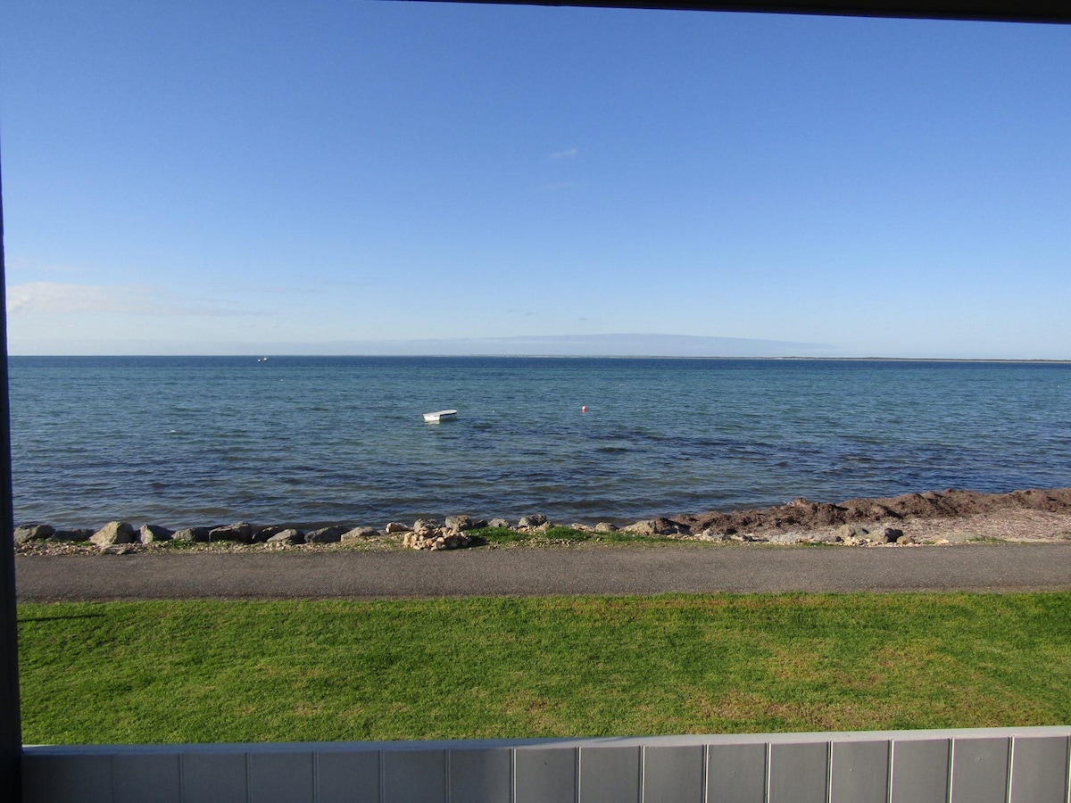 A clear view of the tranquil bay is presented, showcasing gentle waves lapping against the shore. The grassy beachfront is bordered by a walking path, while distant boats can be seen on the water. The sky is bright blue, enhancing the serene landscape.