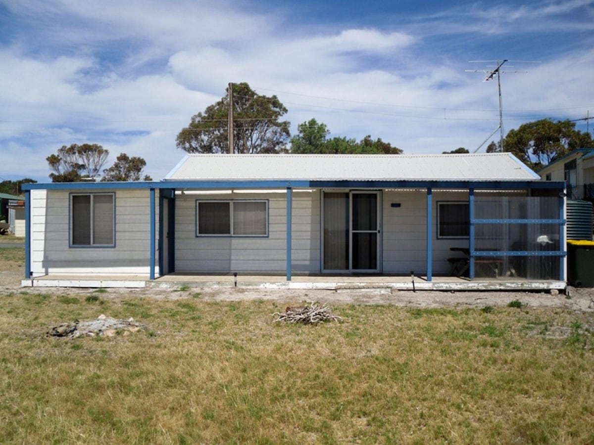 The exterior of a single-story beach shack is depicted, featuring a light-colored facade with blue trim. A covered verandah runs along the front, revealing a set of glass doors, while windows provide views of the surrounding grassy area.