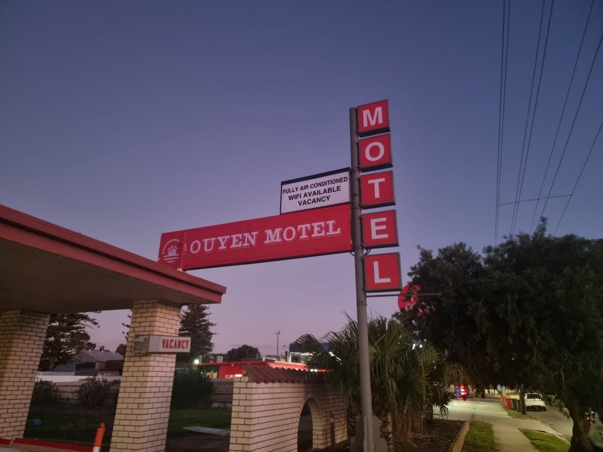 The exterior of Ouyen Motel is displayed at dusk, showcasing a prominent sign with the word 'VACANCY' illuminated. The brick structure, featuring a covered entryway, is set against a hazy twilight sky, with palm trees and surrounding greenery visible nearby.