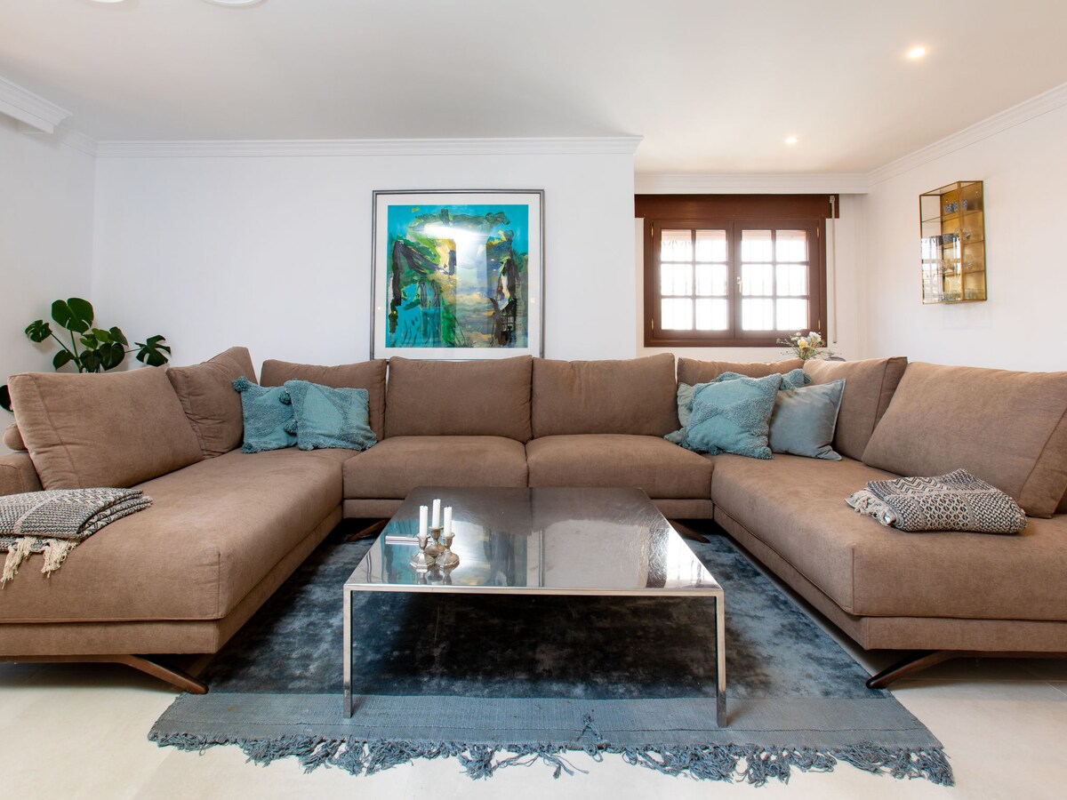 A spacious living room features a large brown sectional sofa adorned with soft throw pillows. A central glass coffee table sits atop a decorative blue area rug. Natural light enters through multiple windows, complementing the modern artwork displayed on the wall.