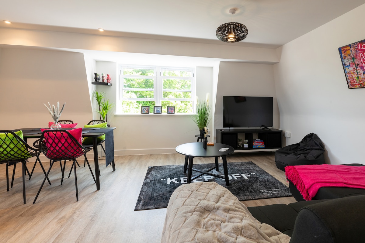 A modern living space showcases a cozy seating area with a dark sofa and a colorful throw. A circular black coffee table sits on a patterned rug. A dining area features black chairs at a dark table, with light streaming in through a large window adorned with indoor plants.