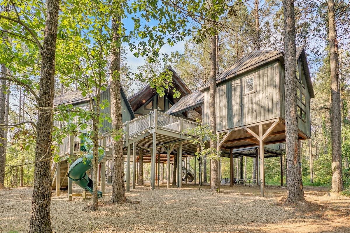 The unique treehouse design features multiple pods elevated above the ground, surrounded by tall trees. A slide is visible on the left side, complementing the outdoor space beneath the structure with seating options among the gravel. Natural light filters through the leaves, enhancing the setting.