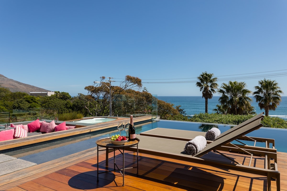 An outdoor terrace features a pool overlooking the ocean, complemented by palm trees and lush greenery. Comfortable lounge chairs are positioned beside a small table holding a bottle of champagne and fresh fruit. The clear blue sky enhances the tranquil setting.