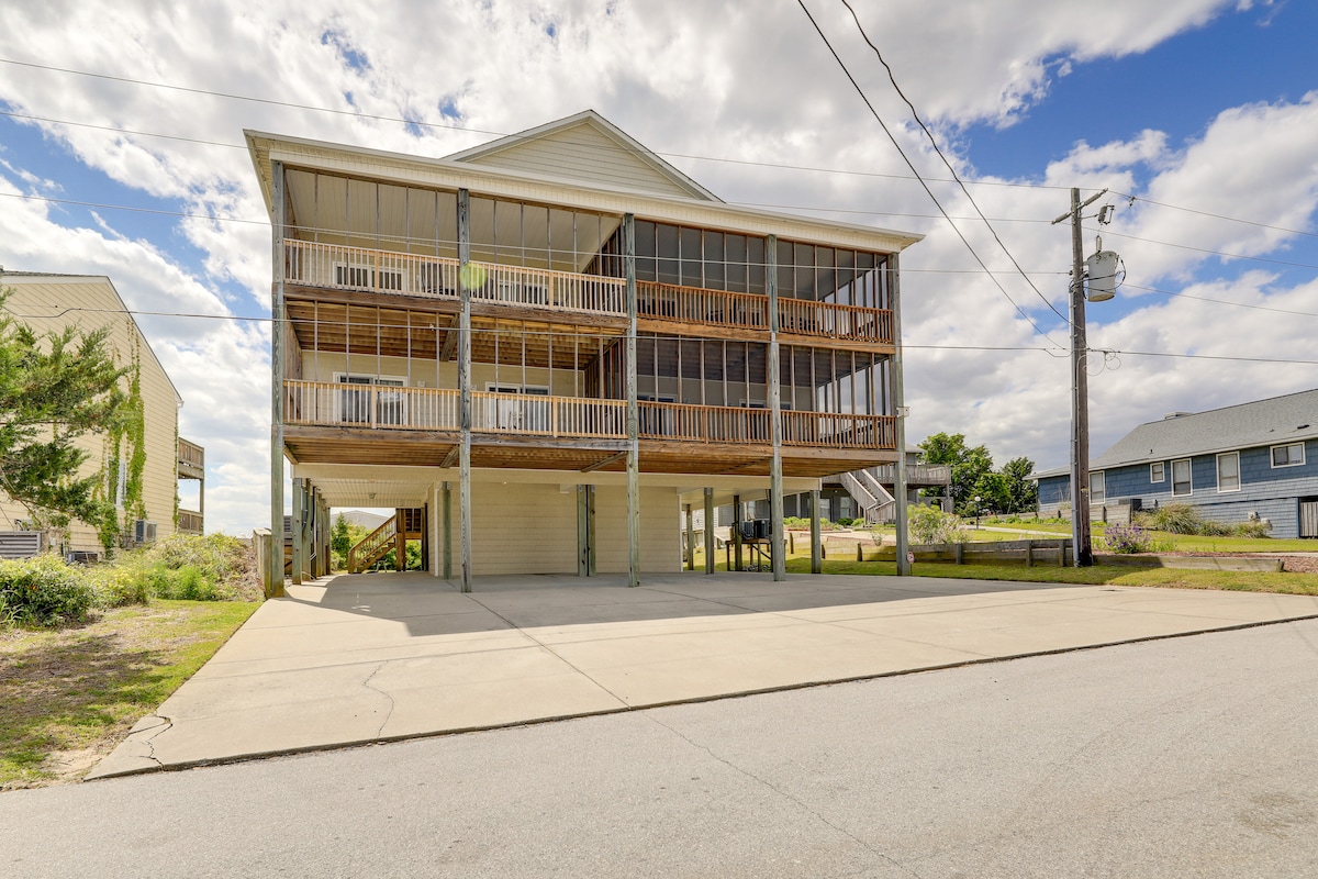 A multi-level townhome with a spacious exterior is shown, featuring screened balconies on the upper levels. The property is elevated, providing a parking lot underneath. Surrounding greenery and open sky are visible, emphasizing the inviting coastal setting.