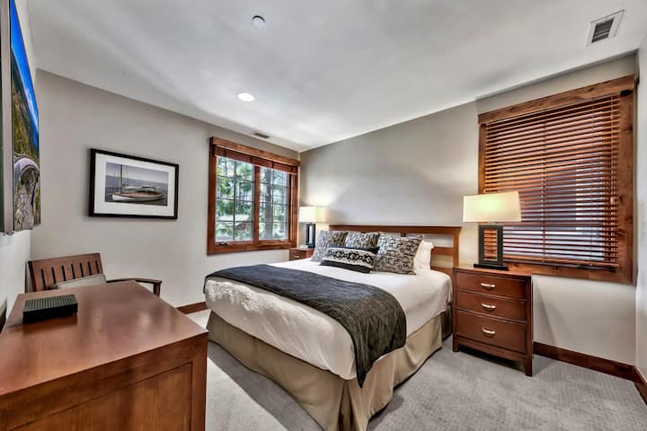 Bedroom with wooden bed frame, patterned throw pillows, two nightstands with lamps, and wood-trimmed windows.