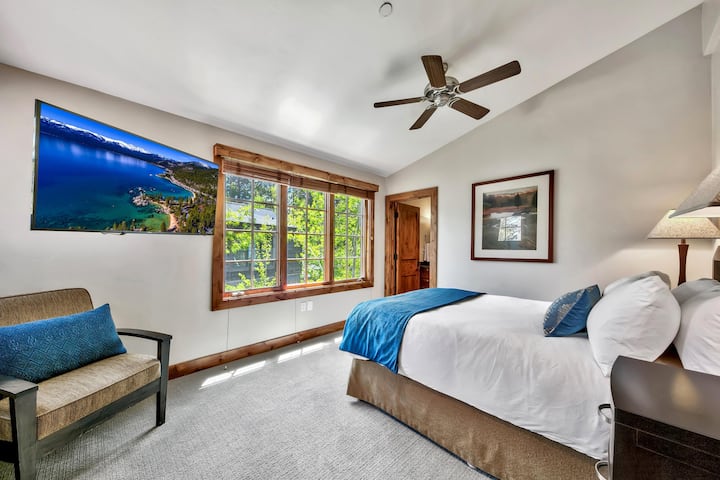 Bedroom with vaulted ceiling, wall-mounted TV, large wood-trimmed window, neutral carpet, and blue-accented bedding.