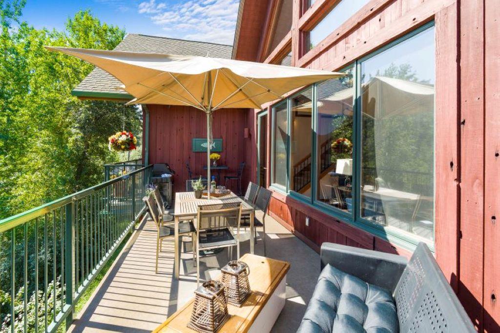 A furnished balcony features a large umbrella providing shade over a wooden dining table with multiple chairs. The area includes a comfortable seating arrangement with cushions and decorative lanterns. Lush greenery is visible in the background, enhancing the outdoor space.