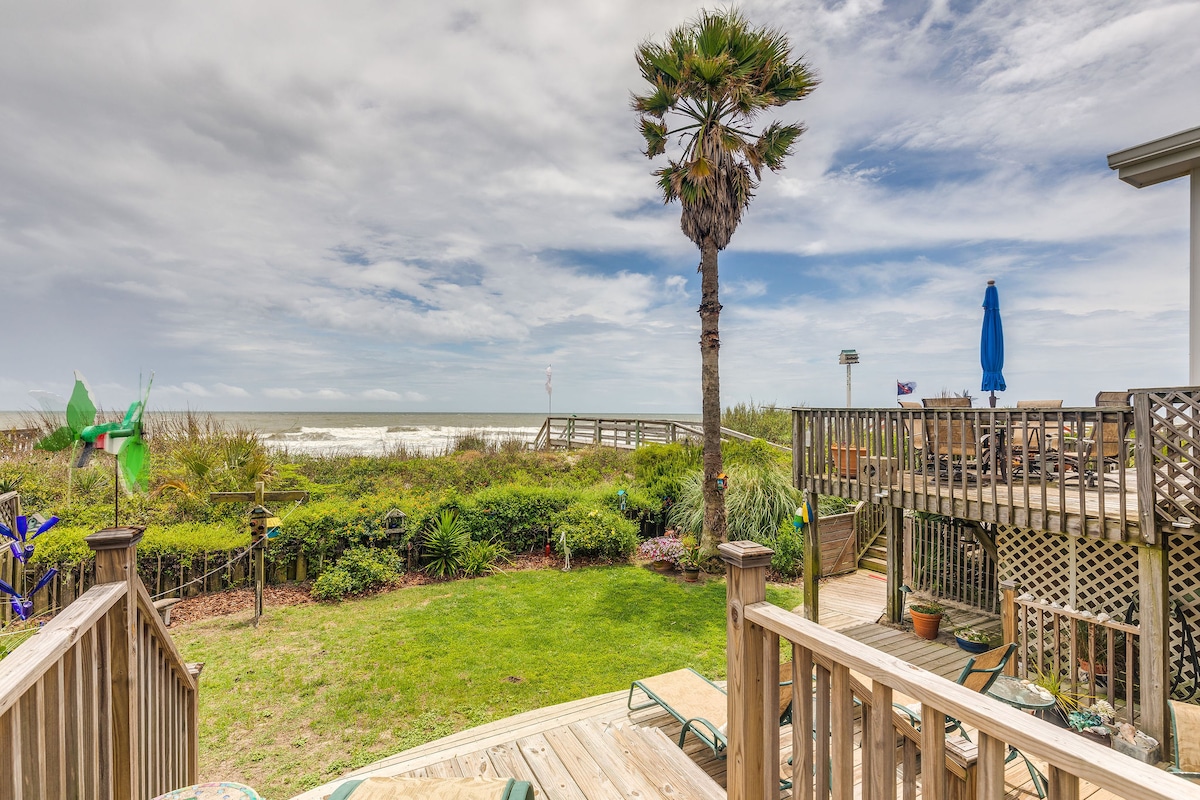 A spacious outdoor area features wooden decking that leads to a well-maintained lawn, framed by tropical landscaping. A palm tree stands prominently, while beach access is visible in the background, showcasing calming ocean views under a partially cloudy sky.