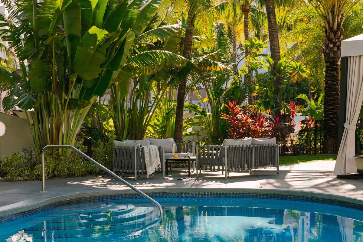 A serene pool area is surrounded by lush tropical plants and palm trees. Comfortable seating featuring grey wooden accents and light-colored cushions is positioned beside the pool. A small table sits between the seating, offering a place for refreshments.