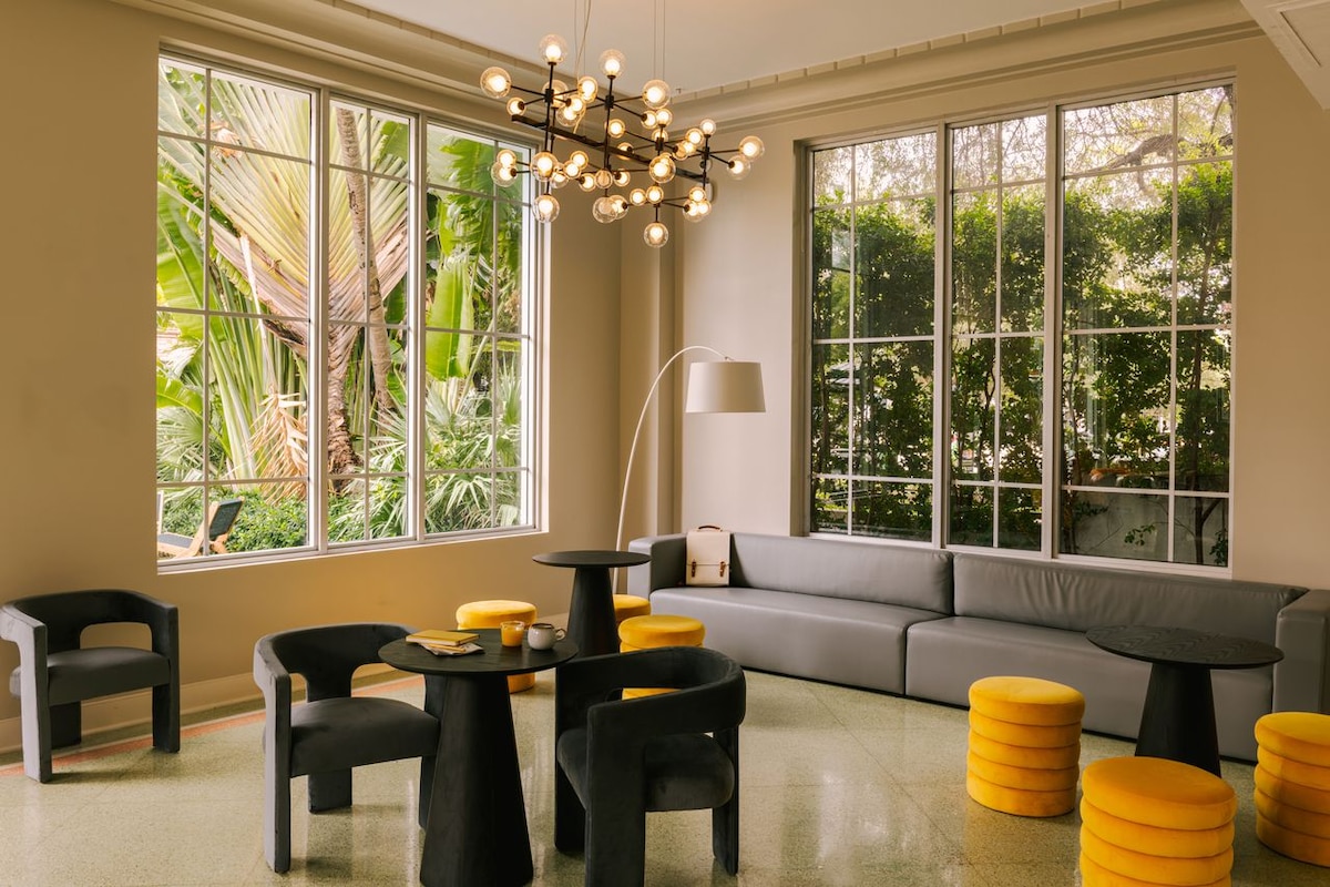 A lounge area features multiple black tables and chairs, complemented by bright yellow stools. Large windows allow natural light to illuminate the space while offering views of lush greenery outside. A stylish chandelier hangs above, adding a modern touch to the room.
