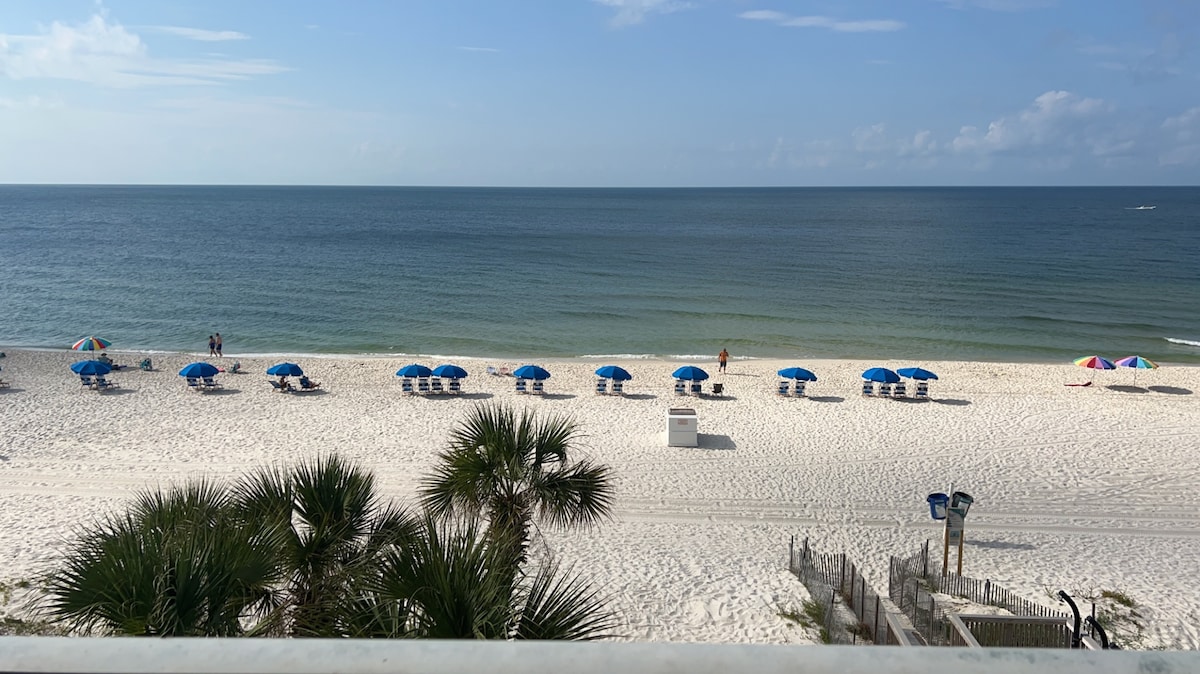 The image captures a serene beach view with white sand and a calm ocean. Beach umbrellas in shades of blue are arranged in neat rows over lounge chairs, while palm fronds are visible in the foreground. A subtle horizon merges the ocean with the clear sky.